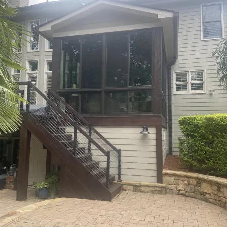 Modern house exterior with dark wood staircase, large windows, beige siding, and stone retaining wall under cloudy sky.