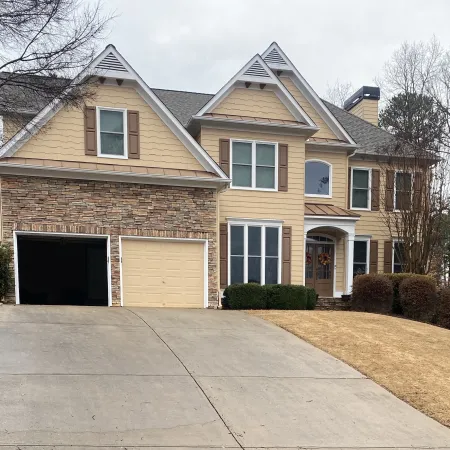 Two-story beige house with stone facade, double garage, large driveway, and landscaped front yard with bushes and trees