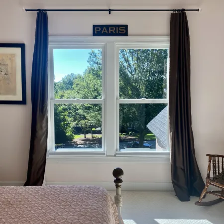 Bedroom window with black curtains framing a bright green outdoor view, pink bedspread, and wooden rocking chair.