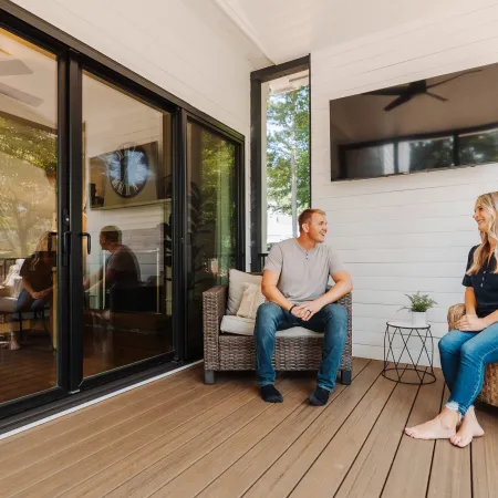 Couple sitting and talking on a modern wooden deck with wicker furniture and a mounted TV indoors.