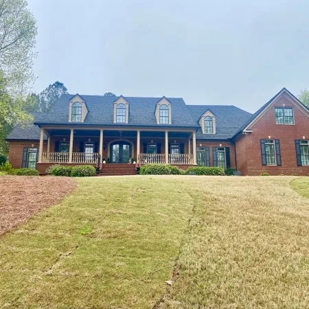 Large brick house with dormer windows, a wooden porch, and a landscaped lawn surrounded by trees under a clear sky.