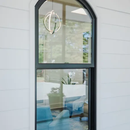 Arched black-framed window on white siding reflects interior with blue chairs and modern light fixture.