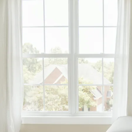A pair of white double hung Infinity windows with white curtains overlooking the neighborhood with a red brick house visible