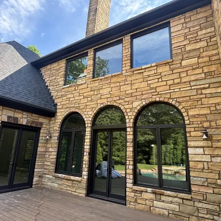 Stone house exterior with arched black-framed windows and double doors opening to wooden deck under blue sky.