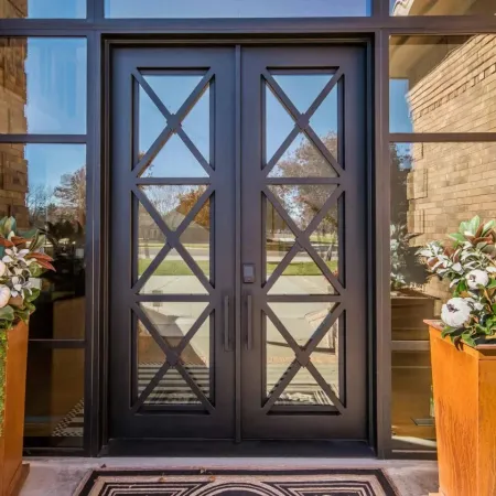 Black metal double front doors with glass panels and decorative X patterns, flanked by large planters with white flowers.