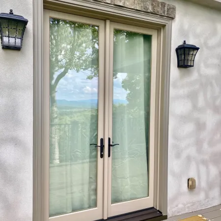 Beige-framed glass French doors with black handles, flanked by two exterior wall lanterns on a stucco home.