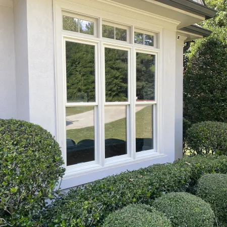 The front of a house with 3 white double hung windows with round and square shaped bushes under the windows