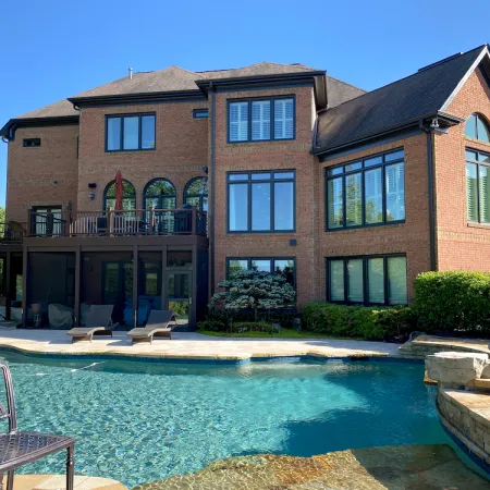 Large brick house with multiple windows overlooks a backyard pool and stone hot tub surrounded by greenery