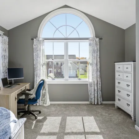 a bedroom with a desk, dresser, twin bed, and a large fiberglass round top window overlooking the neighborhood