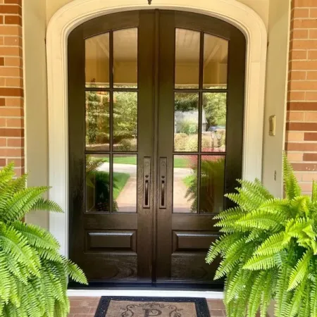 Elegant double black front doors with glass panels flanked by lush green ferns and a personalized doormat on brick floor.