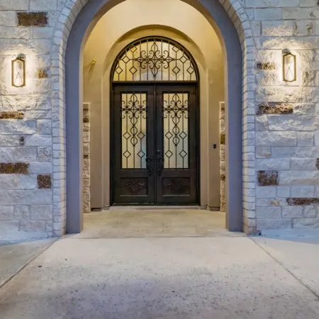 Stone archway entrance with decorative wrought iron double doors illuminated by wall sconces at dusk