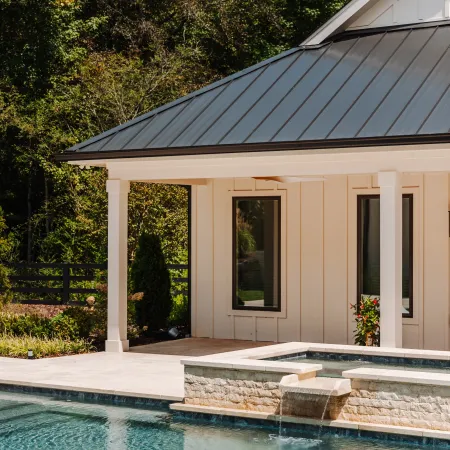 Modern backyard pool with stone waterfall feature and covered patio against lush trees in sunlight.