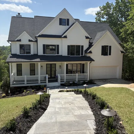 Large white two-story house with black shutters, front porch, stone pathway, and landscaped yard under a blue sky.