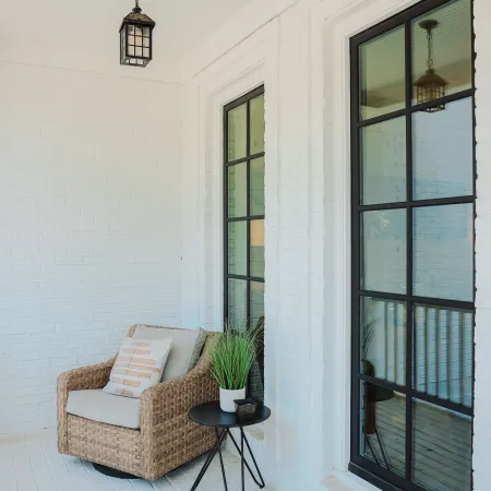 Cozy wicker chair with cushions on a white porch next to black-framed windows and a small round table with a plant.