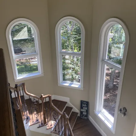 Sunny stairwell with wooden steps, ornate metal railings, and three tall arched windows overlooking a green yard.