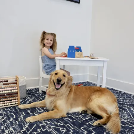 Smiling girl sitting at a white table playing with toys while a happy golden retriever lies on a blue patterned rug.
