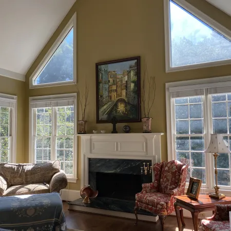 Bright living room with tall windows, two patterned armchairs, a sofa, fireplace, and wall art.
