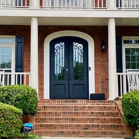 Brick house with white columns and porch railings and arched black iron double front doors with glass