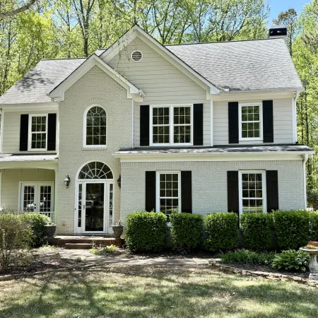 Two-story white house with black shutters surrounded by trees and bushes in a sunny yard.