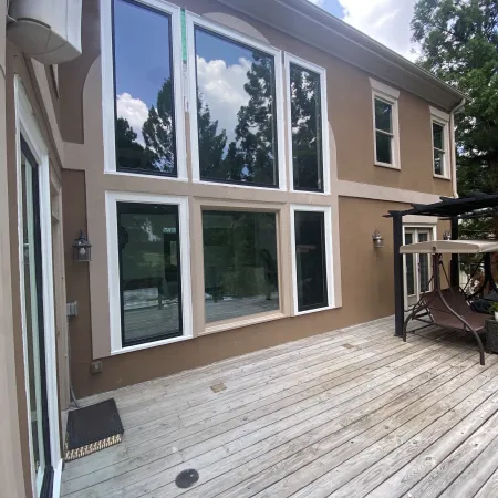 Large windows reflect trees and sky on the exterior wall of a beige house next to a wooden deck and patio swing.