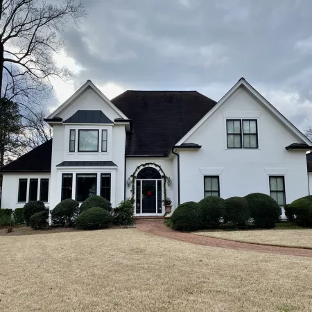 Large white two-story house with dark roof, front shrubs, brick walkway, and barren trees under cloudy sky