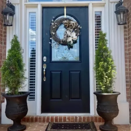 Black front door with decorative glass window and floral wreath, flanked by potted evergreen shrubs and brick steps.