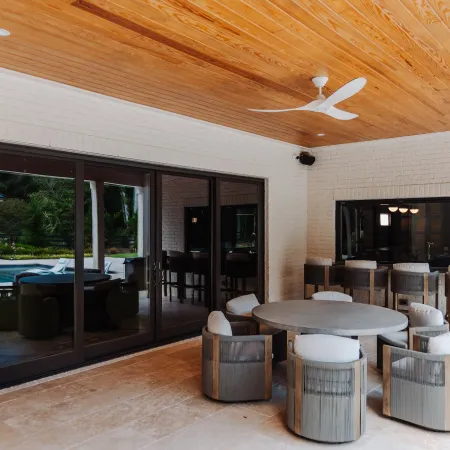 Modern outdoor patio with round dining table, cushioned chairs, wooden ceiling, ceiling fan, and large sliding glass doors.