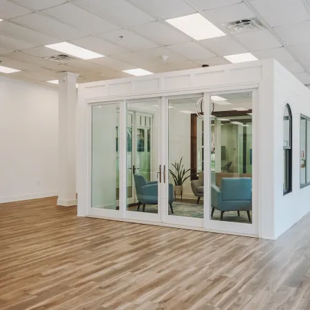 Modern office space with wood flooring, white walls, and glass-walled meeting room with chairs and plant.
