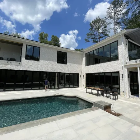 Modern white two-story house with large windows, outdoor pool, and patio under a blue sky with clouds.