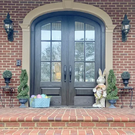 Brick house entrance with double wooden doors, Easter decorations including bunny and pastel eggs, and topiary plants.