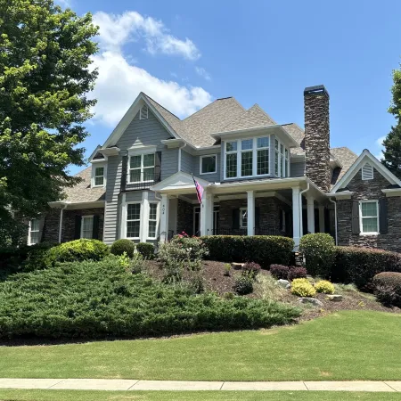 Two-story suburban house with gray siding, stone accents, large windows, and landscaped yard under blue sky.