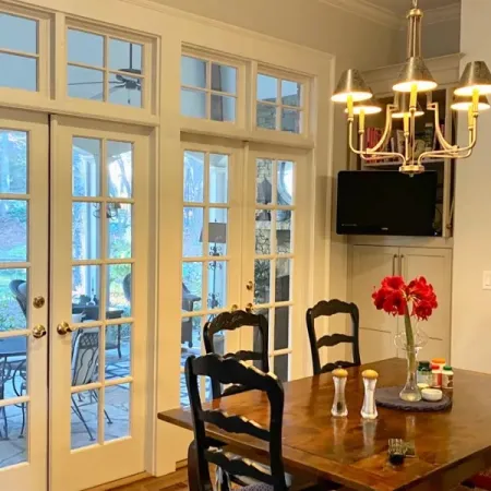 Cozy dining area with wooden table, black chairs, red flower centerpiece, chandelier, and glass French doors.