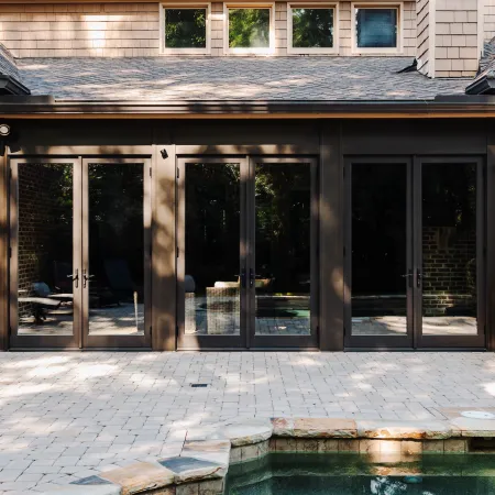 Modern home exterior with large glass doors opening to a patio and pool area in sunlight.