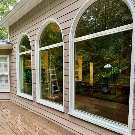Three large arched windows with white trim reflect surrounding trees on a beige house with a wooden deck.