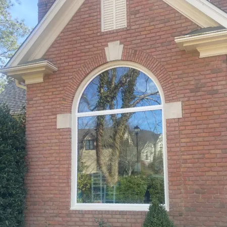 Red brick house front with arched window and door, surrounded by shrubs and a small bush against clear sky.