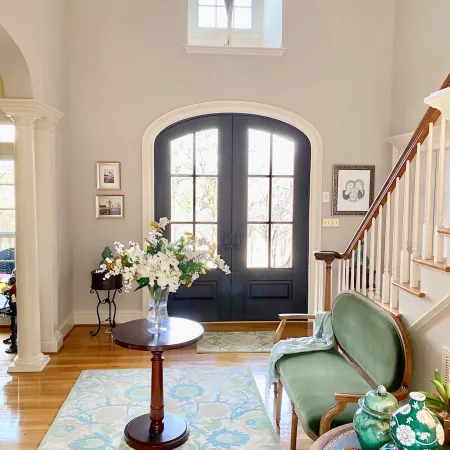 Bright entryway with double black doors, floral centerpiece on table, green velvet loveseat, and decorative ceramics.