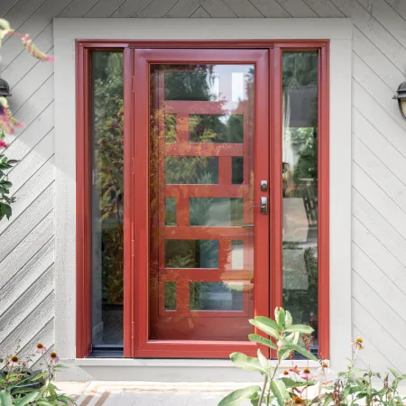Modern red front door with glass panels and sidelights framed by white siding and surrounded by garden plants.