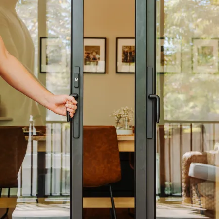 Person opening black-framed glass sliding door to a cozy indoor dining area with chairs and framed photos.