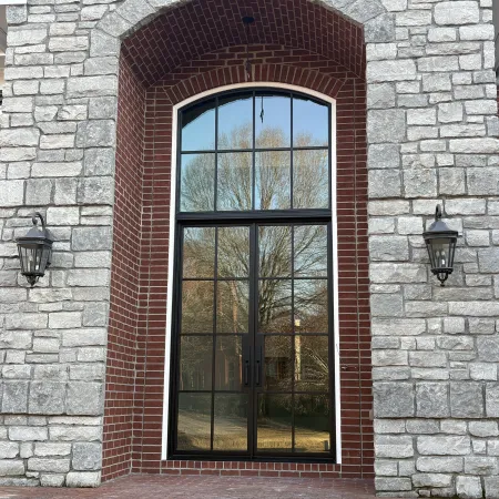 Arched black metal framed glass door with brick trim set in stone wall reflecting outdoor trees and lawn.