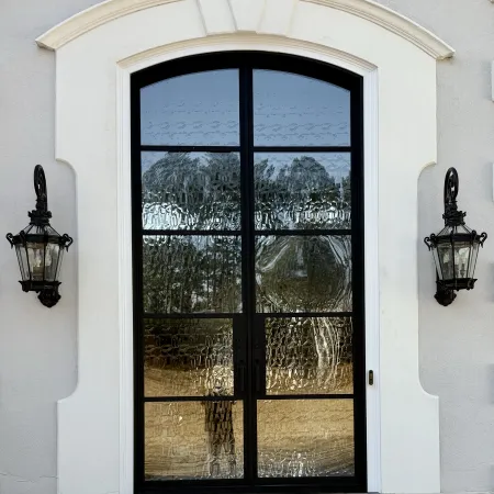 Elegant black-framed arched glass door with textured panes and two vintage lanterns on a light facade.