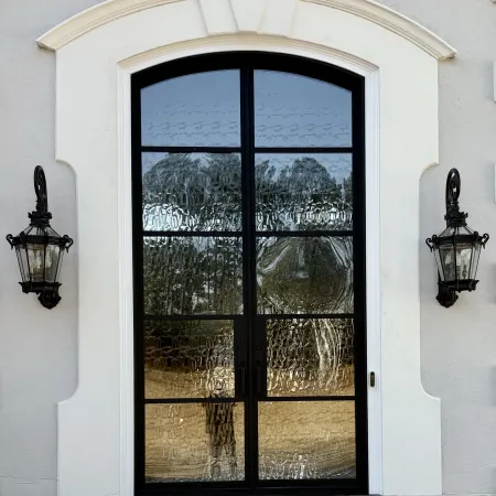 Elegant black iron arched glass door with textured panels beside two decorative black lanterns on white facade.