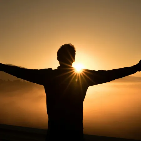 Silhouette of person with arms outstretched facing sunrise over foggy hills and valleys.