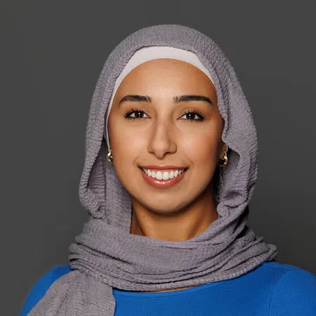 Smiling woman wearing a gray hijab and blue top against a dark gray background.