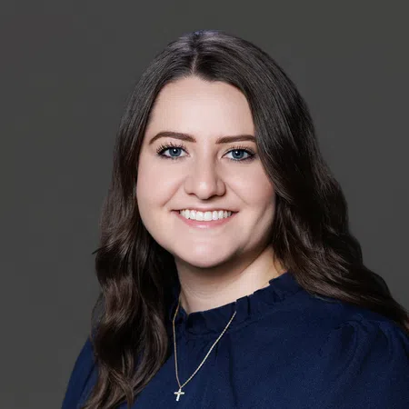 Smiling woman with long brown hair, blue eyes, wearing a navy top and a cross necklace against a gray background