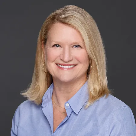 Smiling middle-aged woman with blonde hair wearing a light blue shirt and gold hoop earrings against gray background