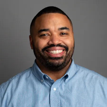 Smiling man with beard wearing a light blue button-up shirt against a plain gray background