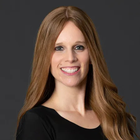 Smiling woman with long light brown hair wearing black top against dark gray background.