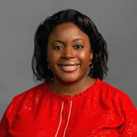 Smiling woman with dark hair wearing red blouse and hoop earrings against a neutral gray background