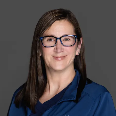 Smiling woman with glasses wearing a blue medical scrub top against a gray background