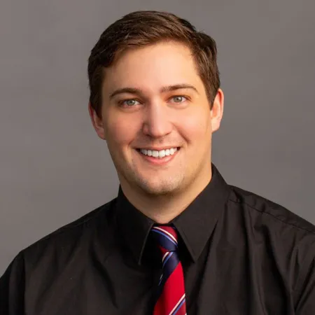 Professional headshot of a smiling man wearing a black shirt and red striped tie against gray background